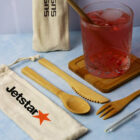 Reusable bamboo utensils, including a fork, knife, spoon, straw, and a cleaning brush, are displayed with two branded cloth pouches labeled Jetstar. A pink drink in a glass is on a wooden coaster. The background is a light blue surface.