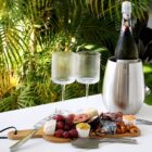 A bottle of sparkling wine rests in a metallic cooler on a table. Two empty wine glasses are beside a charcuterie board filled with cheeses, grapes, nuts, and spreads. Lush green plants serve as the backdrop.
