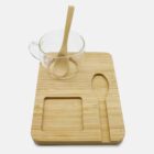 A glass cup with a wooden spoon inside sits on a rectangular bamboo tray. The tray has cutouts for the spoon and cup, as well as an additional square section. The background is plain white.