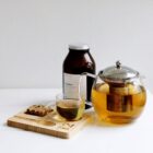A glass teapot filled with tea sits on a white surface alongside a clear teacup. Next to them, a dark jar labeled Grand blend is visible. A wooden tray holds a tea mug-shaped coaster and a piece of granola.