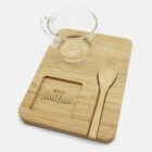 A clear glass cup and a wooden spoon are placed on a rectangular wooden board. The board has an indent featuring the Tim Tam logo, designed for holding biscuits. The background is plain white.