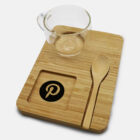 A bamboo tray with a transparent glass cup, a wooden spoon, and a square indentation featuring a stylized letter P logo. The tray has a minimalist design on a white background.
