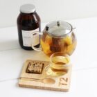 Glass teapot and cup filled with golden tea on a wooden tray with a spoon and a cookie. A brown jar labeled Coastal Blend White Tea is in the background on a white surface.