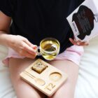 A person sits on a white surface, holding a clear mug of tea and a book. A wooden tray with a cookie, a spoon, and a round space for the mug rests on their lap. Theyre wearing a black top and pink shorts.