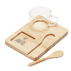 A transparent glass cup sits on a wooden tray with a carved slot next to it. A wooden spoon rests beside the tray, which features an engraved design. The bamboo textures of the tray and spoon are evident.