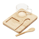 A clear glass mug sits on a rectangular wooden tray with grooves for a perfect fit. A small wooden spoon rests beside the tray. The tray has a minimalistic design with a slot for the spoon and another curved groove.
