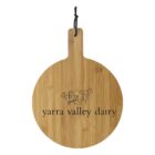 A round wooden cutting board with a handle and a black hanging cord. It features a line drawing of a cow and the text yarra valley dairy engraved on its surface.