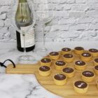 A round wooden board with small chocolate tarts topped with sea salt rests on a marble surface. In the background, two wine glasses and two wine bottles stand in front of a white brick wall.