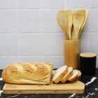 A loaf of bread partially sliced on a wooden cutting board with Disney engraved. Behind it, theres a holder with wooden utensils and a black container with a wooden lid. The background has white tiles.
