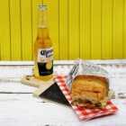 A bottle of Corona Extra beer is on a wooden board next to a sandwich wrapped in newspaper print paper with a red and white checkered napkin underneath. The background is a bright yellow wall.