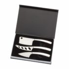 A set of three cheese knives with stainless steel blades and white handles, neatly arranged in a black box. The set includes a cleaver-style knife, a pronged knife, and a rounded cheese spreader.