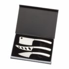 A set of three cheese knives in a black box with a lid. The knives have stainless steel handles and distinct blade shapes: one with holes, one wide-bladed, and one pointed, designed for different cheese types.