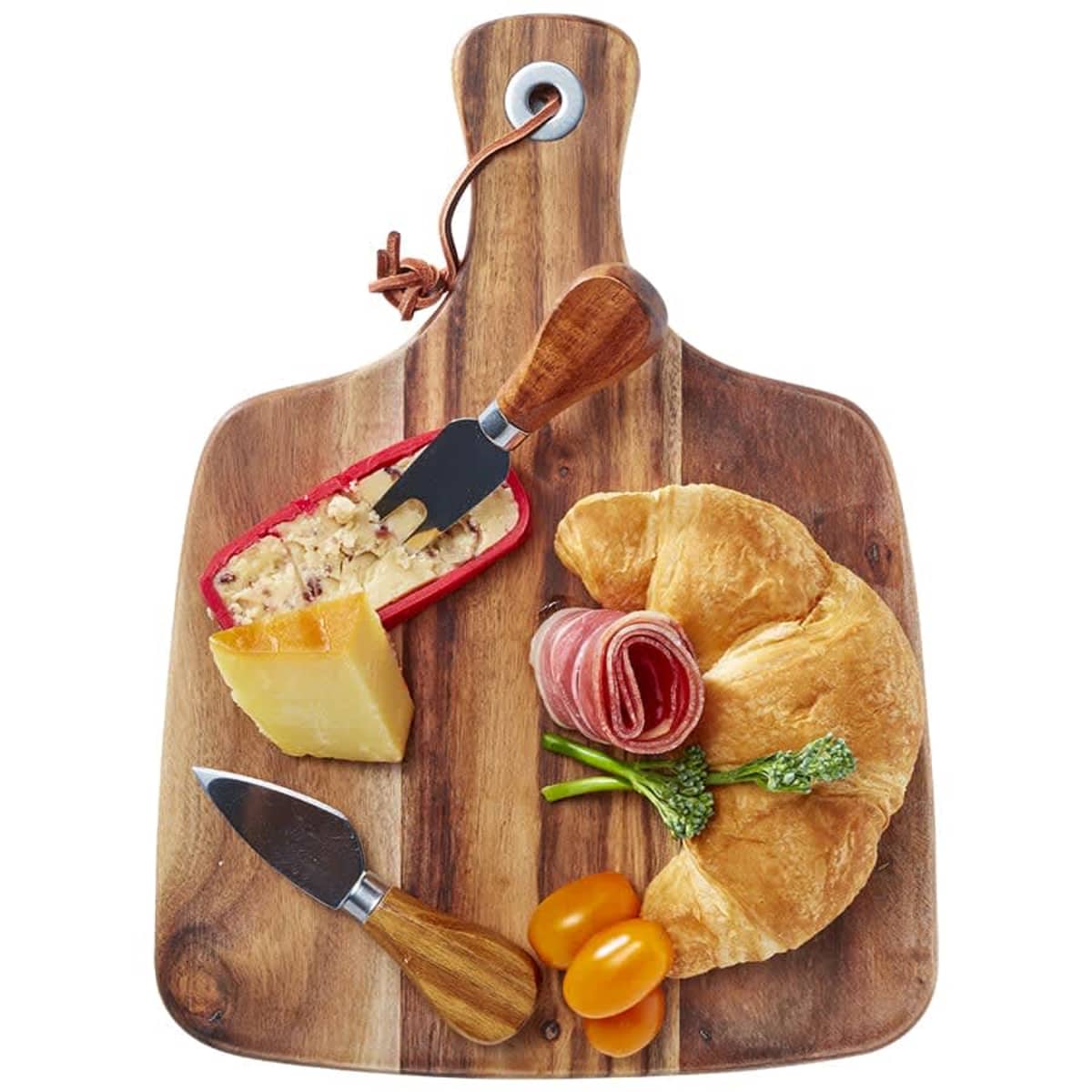 A wooden cutting board holds a cheese wedge, a red cheese block with a fork, slices of cured meat, a croissant, broccoli, and two cherry tomatoes. Two cheese knives with wooden handles are also on the board.
