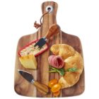 A wooden serving board with a croissant, slices of cured meat, cheese, a leafy vegetable, and two yellow cherry tomatoes. Two cheese knives rest on the board, one inserted into a cheese wedge.