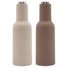 Two modern, cylindrical salt and pepper grinders with matte finishes. One is light beige, and the other is light brown. Both have silver tops. They are positioned side by side against a white background.