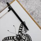 A detailed black and white drawing of a butterfly on a white sheet, clipped to a board. A black pen is placed diagonally across the top left corner of the paper. Intricate swirling patterns are visible in the background.