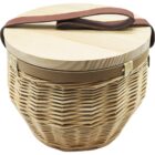 A round wicker basket with a wooden lid and a brown leather strap handle. The basket has a woven texture and a sturdy design. The lid fits snugly, giving it a rustic and practical look.
