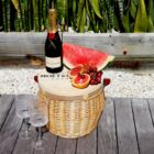 A wicker picnic basket with a bottle of Moët & Chandon champagne, a large watermelon slice, and a bunch of grapes sits on a wooden surface. Three empty wine glasses are beside the basket, with green plants in the background.