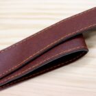 Close-up of a brown leather belt with yellow stitching on a light wooden surface. The belt is folded over itself, showcasing its texture and craftsmanship.