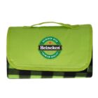 A folded green picnic blanket with a handle, featuring the Heineken logo prominently on the front flap. The blanket has a black and green checkered pattern visible at the bottom.