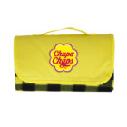 A folded picnic blanket with a yellow flap and a handle, featuring the Chupa Chups logo, rests against a white background. The lower part shows a black and green checkered pattern.