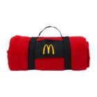 A red blanket rolled up and secured with black straps. In the center is a black patch with a yellow arch logo. A black handle is attached for easy carrying.