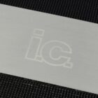A metal surface with the faint outline of the letters ic is placed on a textured black background. The letters are subtly impressed and slightly shiny, blending with the surface.