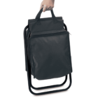 A person holding a black folding chair with a built-in carrying handle and attached bag. The chair has metal legs and a compact design suitable for easy transport.