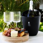 A bottle of sparkling wine in a black cooler with two empty stemmed glasses beside it. Theres a wooden platter with assorted cheeses, meats, nuts, and fruits. Lush green foliage serves as the backdrop.