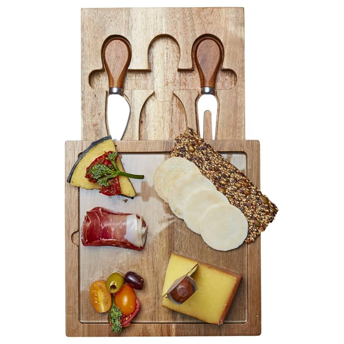 Wooden cheese board with cheeses, sliced meats, and assorted garnishes. Two cheese knives and a fork are nestled in dedicated slots above the board. Various cheese slices, a rolled cured meat, grapes, and cherry tomatoes are arranged on the board.