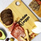 A wooden board with Rockdale & Leggo text features sliced dark bread, salami, prosciutto, breadsticks, Brie cheese, half an avocado, olives, and a slice of seeded bread. The board rests on a textured white surface.