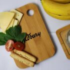 A wooden cutting board with cheese slices, cherry tomato halves, fresh greens, and breadsticks. Two bananas are beside the board on the right, all placed on a white surface.