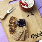 Crackers and seed bread slices arranged on a wooden board, accompanied by a small bowl of olives and a ceramic dish of dip garnished with red chili peppers. A cheese fork is placed nearby. The board has the Carlsberg logo.