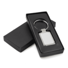 A rectangular silver keychain sits in a black open box with a lid placed underneath. The keychain is neatly positioned, showcasing its reflective metallic surface. The overall presentation is sleek and elegant.