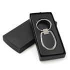 A metal keychain with a modern design is placed in an open black gift box. The keychain has an oval shape and a square connecting piece. The box and keychain are set against a plain white background.