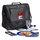 A black insulated picnic bag with Almaty Apple Festival and a red apple logo. In front, a blue plate with food, black-handled cutlery, a clear glass, salt and pepper shakers, a corkscrew, and a folded napkin.