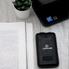 A black leather case branded Lexus lies on a white wooden surface beside an open book. A small green potted plant and a laptop with a visible Intel sticker are placed nearby.