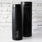 A sleek, black water bottle with the word dusk printed on it stands on a white textured surface. Behind it is a matching black cylindrical box with a white line design. The background is a light-colored brick wall.