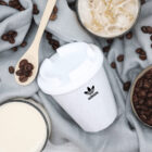 A white cup with a lid featuring an Adidas logo, surrounded by a wooden spoon with coffee beans, two glasses—one with iced coffee and one with a light beige liquid—on a gray fabric background scattered with coffee beans.
