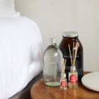 A bedside table with a large brown jar labeled Mayde Tea, a clear bottle labeled podes, and two small diffuser bottles with the Airbnb logo. The corner of a white bed is visible in the background.