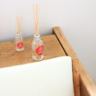 Two small glass bottles with reed diffusers sit on a wooden table. Each bottle has the Airbnb logo on it. The table is next to a cushioned backrest. The background is a plain white wall.