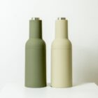 Two sleek, modern salt and pepper grinders stand side by side on a white surface. One grinder is olive green, and the other is beige. Both have a cylindrical shape with stainless steel tops, set against a plain white background.