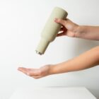 A person dispenses liquid from an upside-down, beige bottle into their open hand. The background is plain white, and the image focuses on the hands and the bottle.