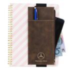 A spiral-bound notebook with diagonal pink and white stripes is shown. A brown leather pocket, embossed with the Mercedes-Benz logo, holds a blue pen and conceals part of a smartphone and some paper currency.