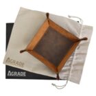 A brown leather tray with raised corners sits atop a beige drawstring bag. Below the bag is a black box. Both the bag and box have the text AGRADE printed on them in white.