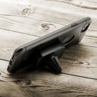A smartphone with a black case is propped up by a phone holder attachment on a wooden surface. The phone holder is designed for mounting on surfaces, providing a stable support for the device.