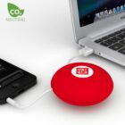 A red circular device with Uniqlo logo is connected via USB cables to a smartphone and a laptop. A CO2 Neutral logo is on the top left. The smartphone is black and the laptop is silver.
