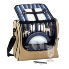 A picnic bag with an open flap reveals cutlery, plates, and cups neatly arranged inside. The interior has a striped blue and white pattern. A shoulder strap is attached to the beige exterior, and a corkscrew and glass are in front.