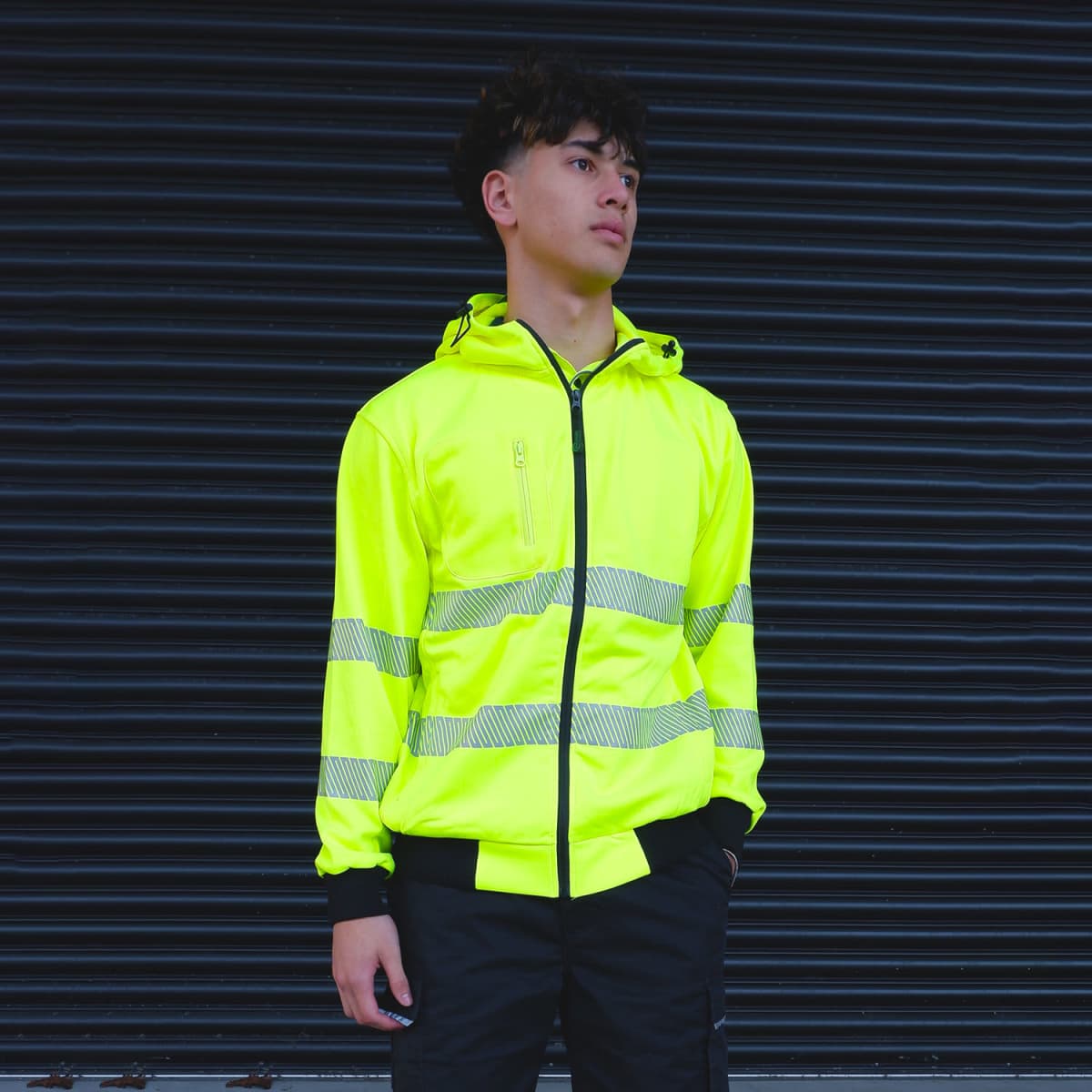 A person wearing a bright yellow reflective jacket stands in front of a dark, ribbed background. The jacket has gray reflective stripes, and the person appears to be looking into the distance with a neutral expression.