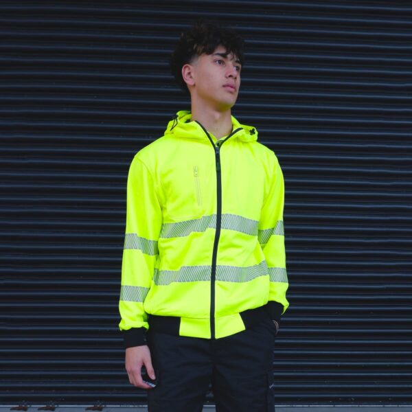 A person stands in front of a dark, ribbed background, wearing a bright yellow reflective jacket with gray stripes and black pants. The jacket has a hood, and the person looks slightly to the side.